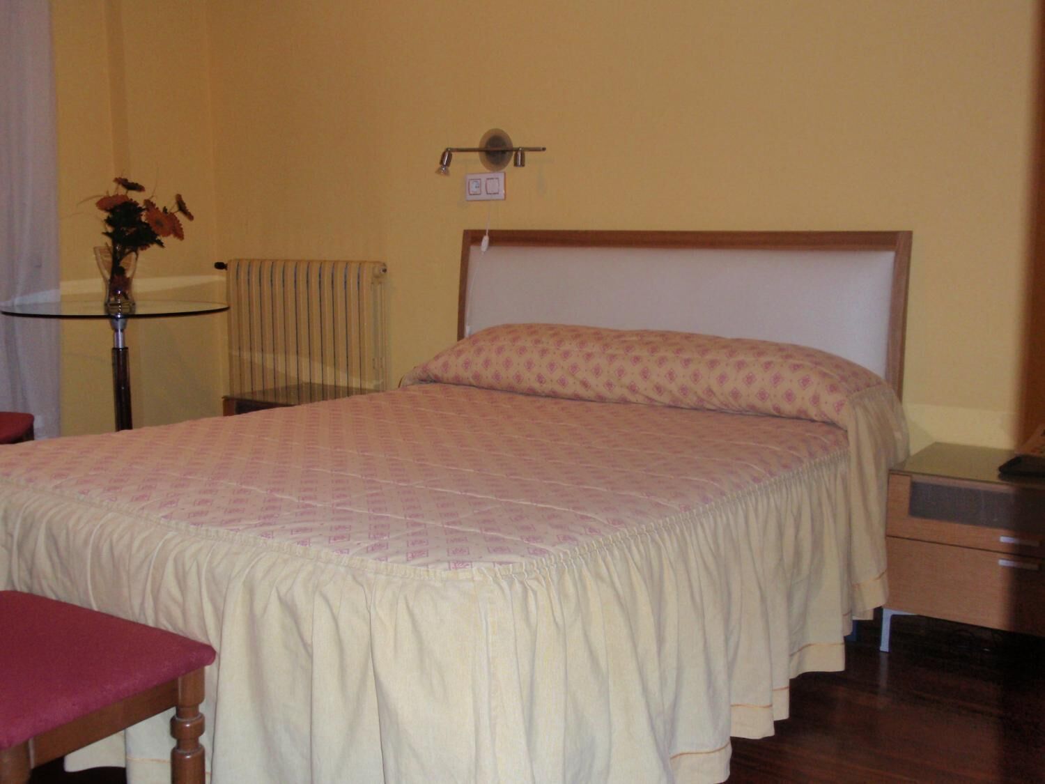A neatly made bed with a patterned bedspread, a wooden nightstand, a small table with a vase, and a radiator.
