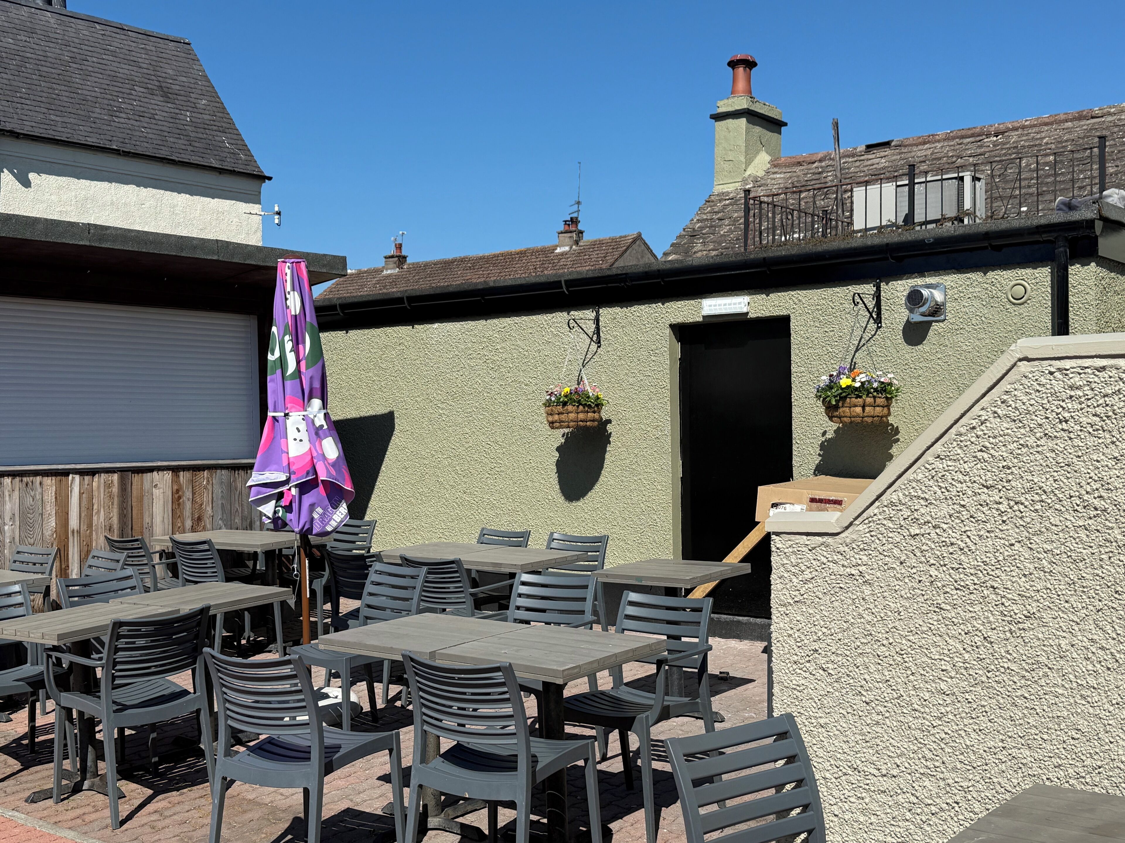 An outdoor patio with tables and chairs, a purple umbrella, and hanging flower baskets.