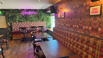 A restaurant interior with wooden tables and chairs, a neon sign, and a mural on the wall.