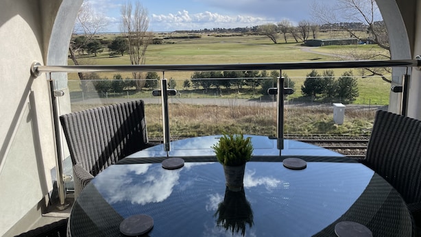 A balcony with a glass railing, a round table with a potted plant, and a view of a golf course. - 19th Hole Hotel (Carnoustie)