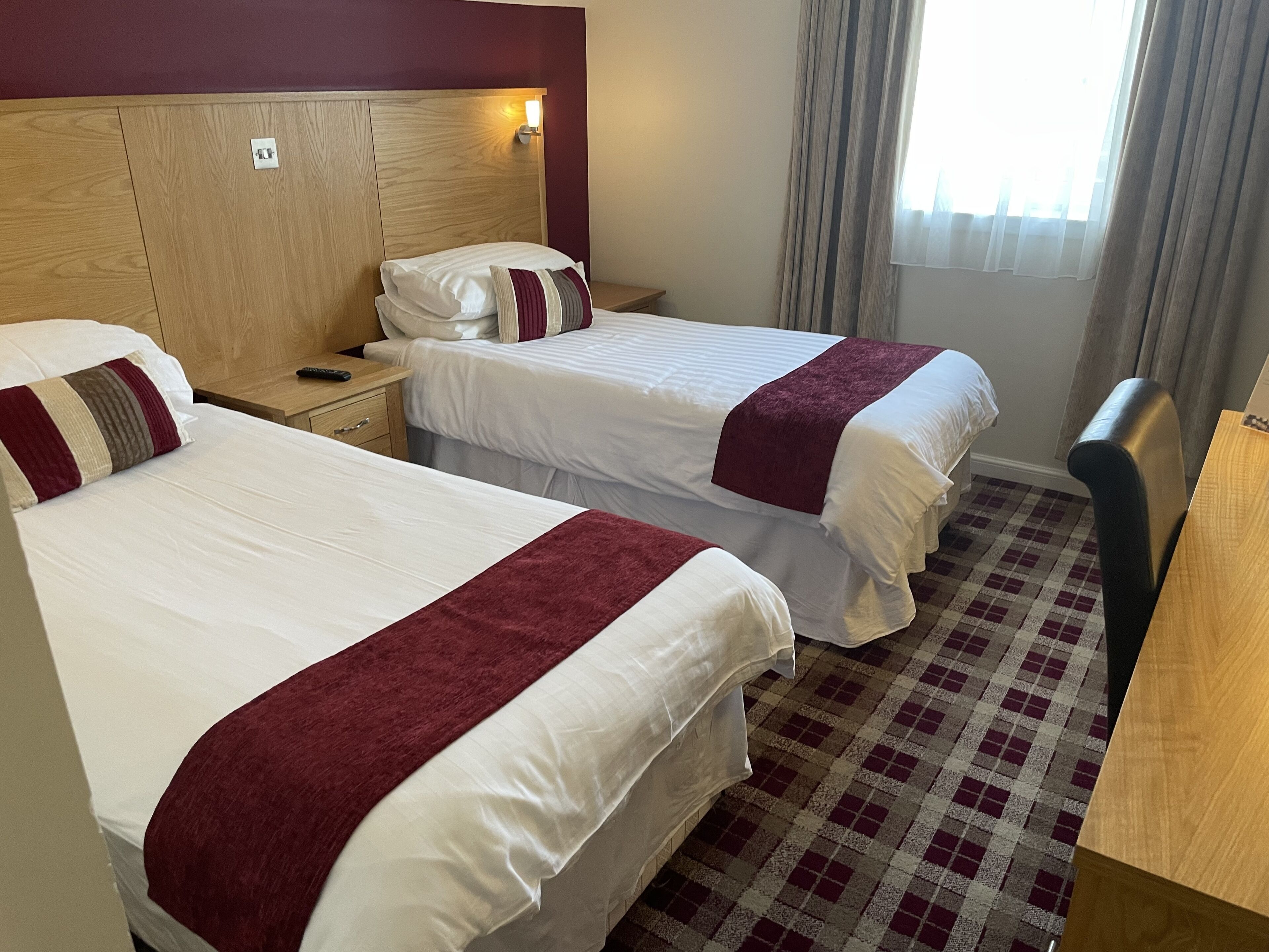 Une chambre d’hôtel avec deux lits, chacun recouvert de draps blancs et d’une couverture rouge. On y trouve une tête de lit en bois, une table de chevet avec une lampe et un sol à carreaux.