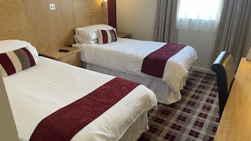 Une chambre d’hôtel avec deux lits, chacun recouvert de draps blancs et d’une couverture rouge. On y trouve une tête de lit en bois, une table de chevet avec une lampe et un sol à carreaux.