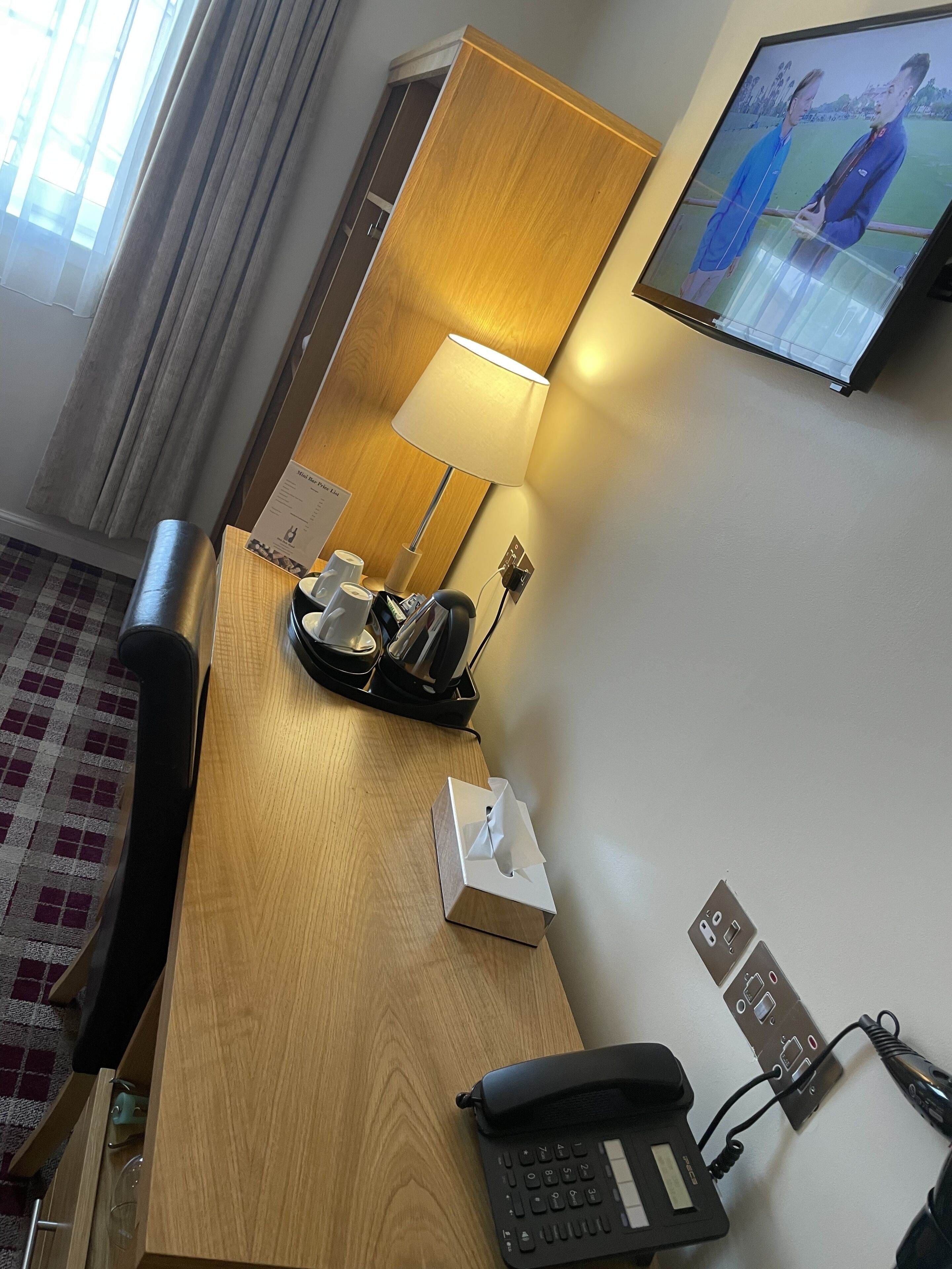 Habitación de hotel con escritorio de madera, teléfono, lámpara, pantalla de televisión y mostrador de recepción.
