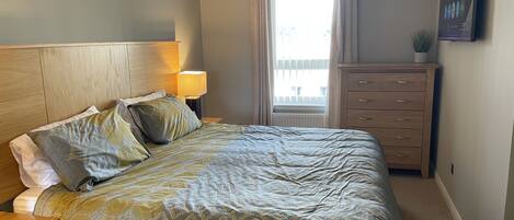 A bedroom with a bed, a nightstand, a dresser, a lamp, and a window with curtains.