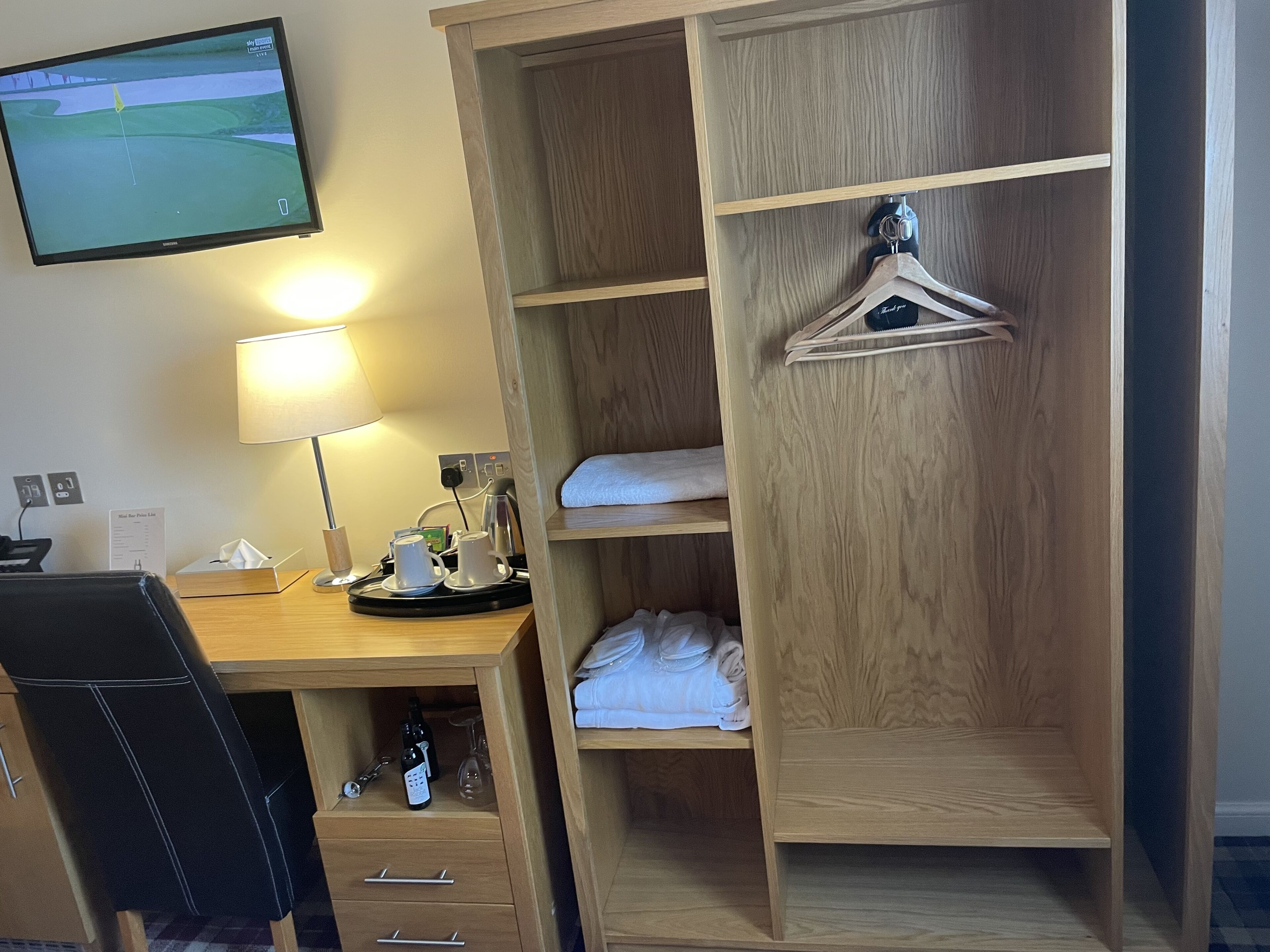 A hotel room with a wooden desk, a TV mounted on the wall, a lamp, a coffee machine, and a wardrobe with hangers.