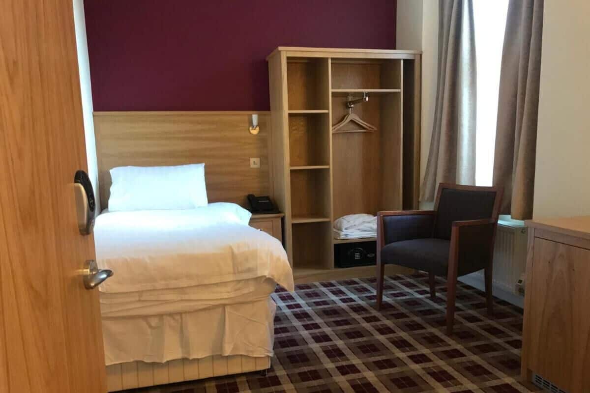 A hotel room with a bed, a wardrobe, a chair, and a check-in desk.