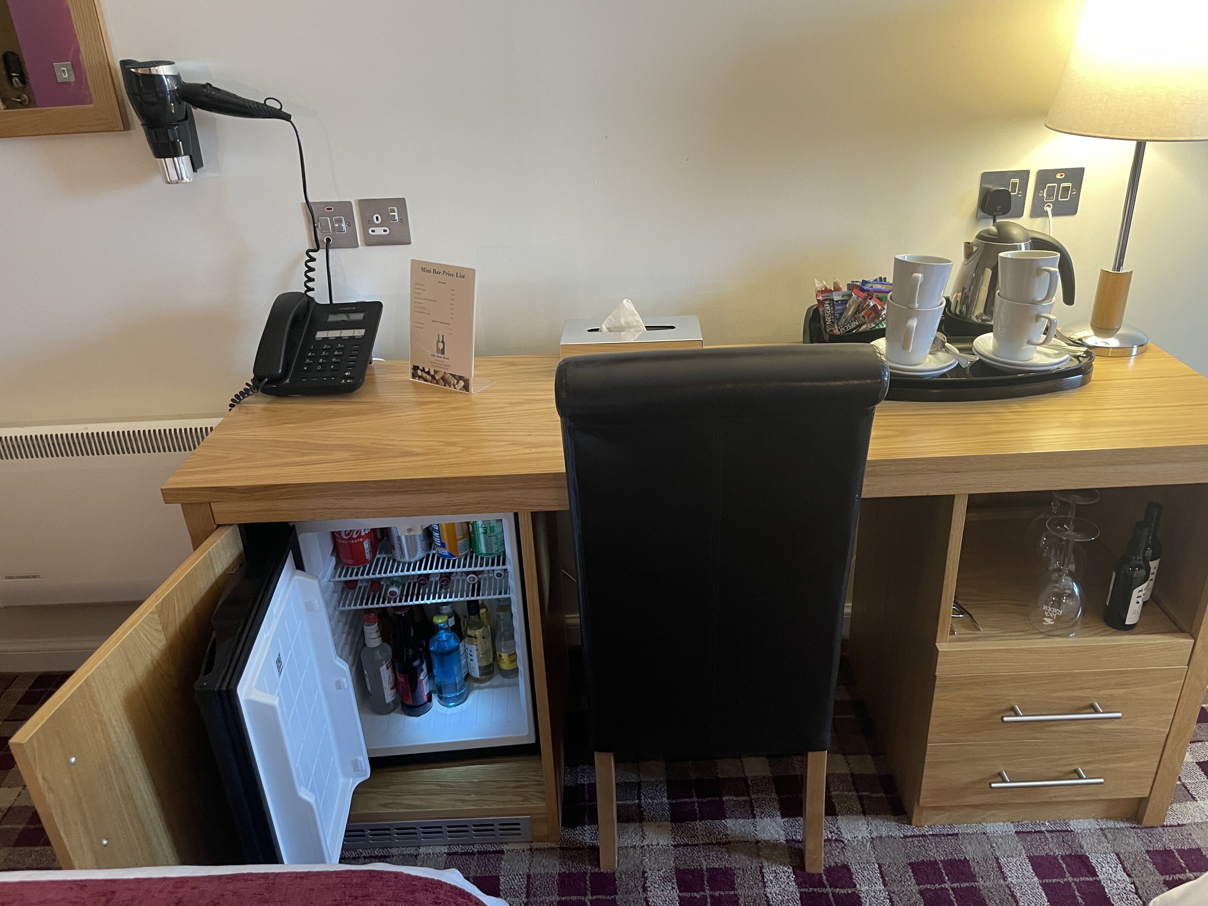A hotel room with a wooden desk, a black office chair, a mini fridge with beverages, a coffee maker, and a lamp.