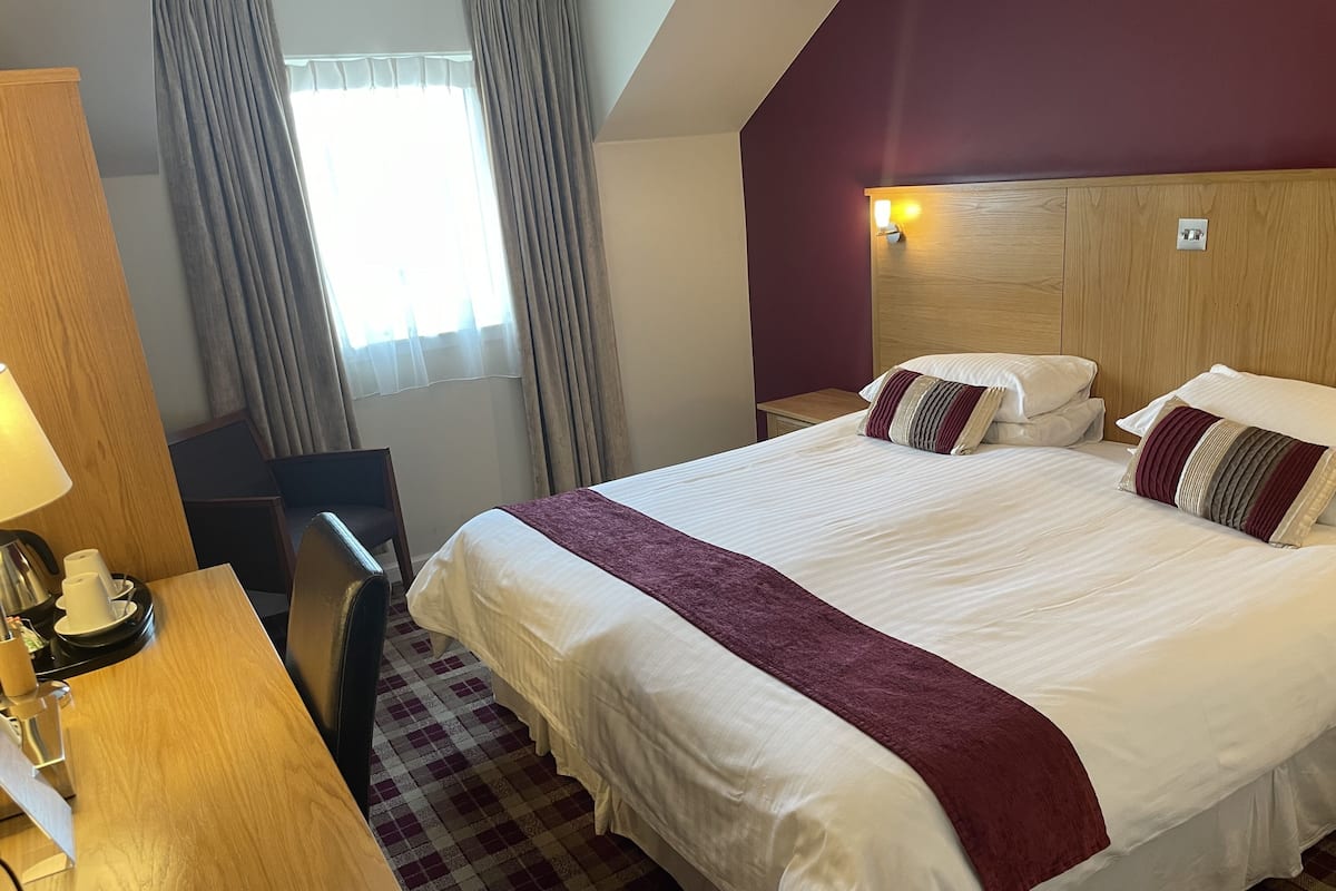 A hotel room with a large bed, a desk with a lamp, a chair, and a check-in counter.