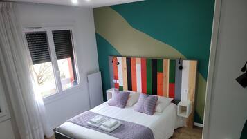 Een netjes opgemaakt bed met kussens, een nachtkastje met lamp, een raam met zonwering en een kleurrijke muur.