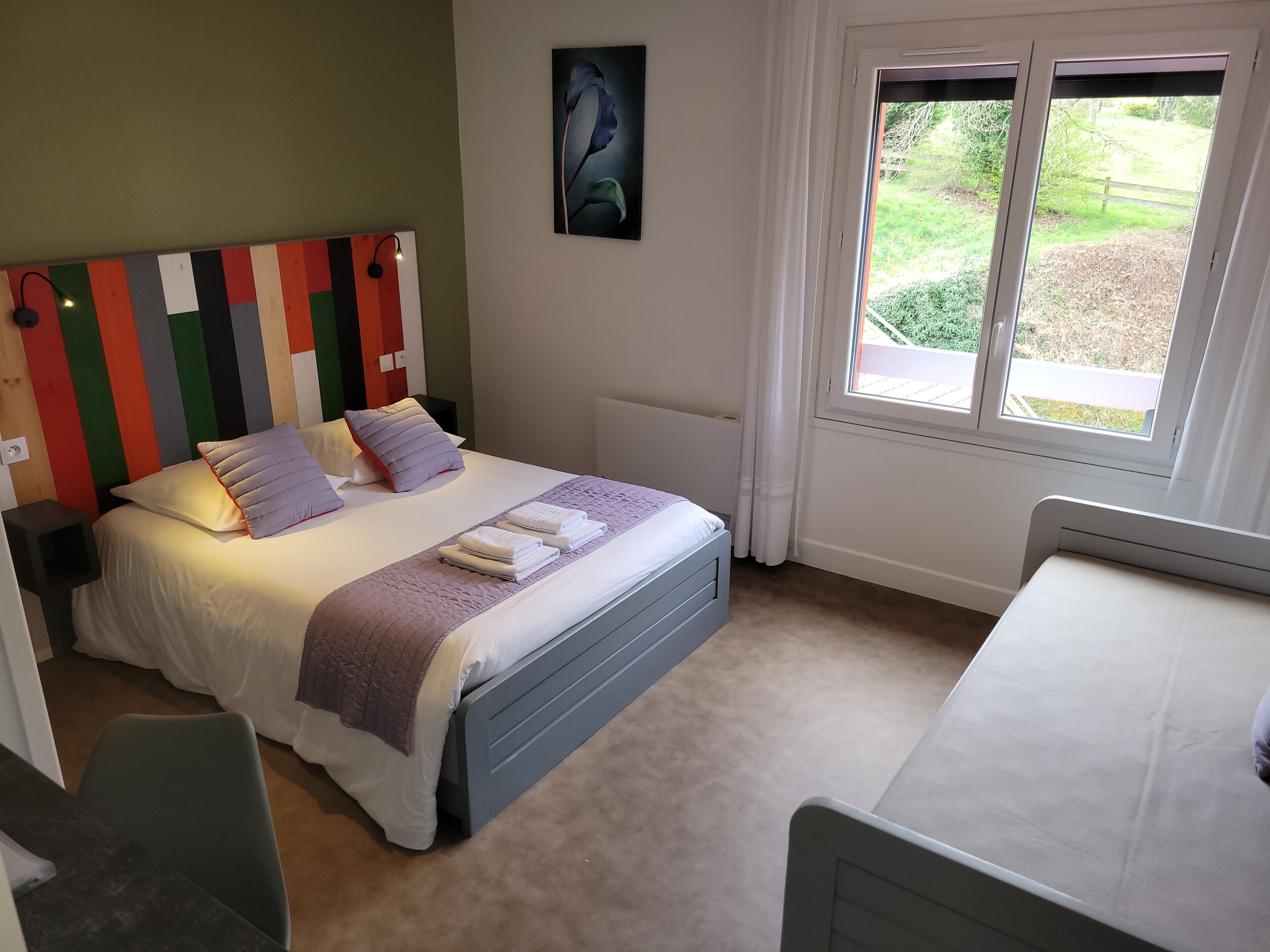 Une chambre à coucher comprenant un lit, des tables de chevet, une fenêtre donnant sur l’extérieur et un mur agrémenté d’une tête de lit colorée.