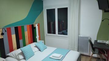 A hotel room with a bed, a desk, a chair, a radiator, and a window with curtains.