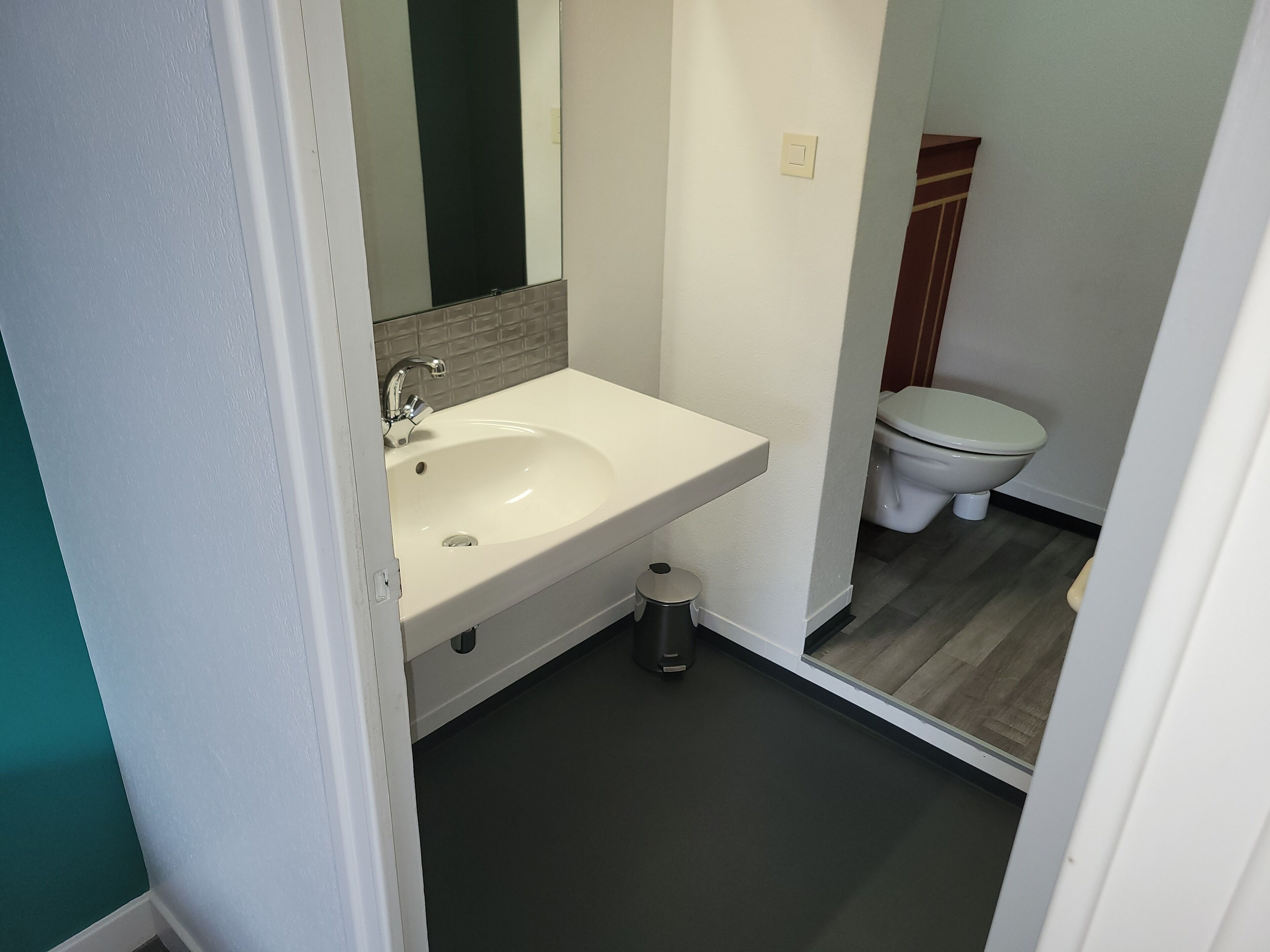 Ein Badezimmer mit einem weißen Waschbecken, einem Spiegel, einer Toilette und einem Mülleimer.