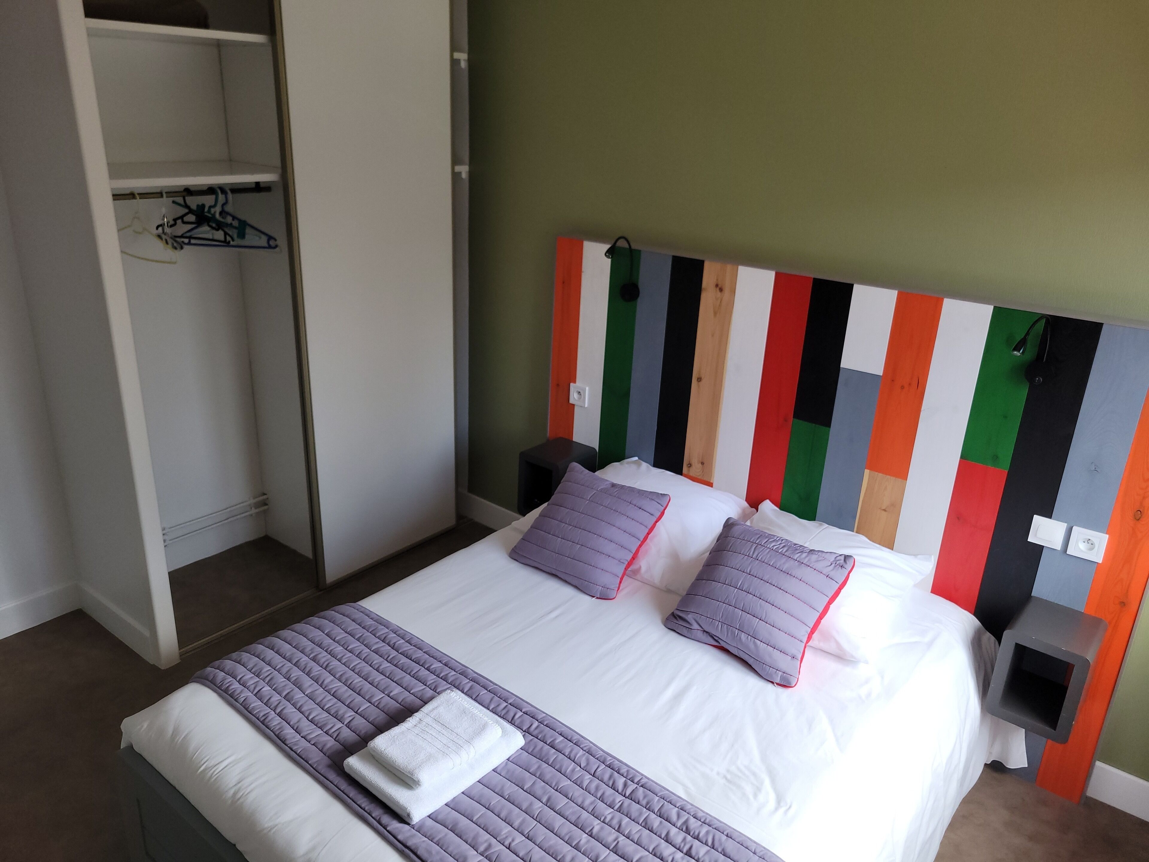 A neatly made bed with a purple quilt, a white pillow, and a folded towel. There is a white wardrobe with a hanging rack and a colorful striped headboard.