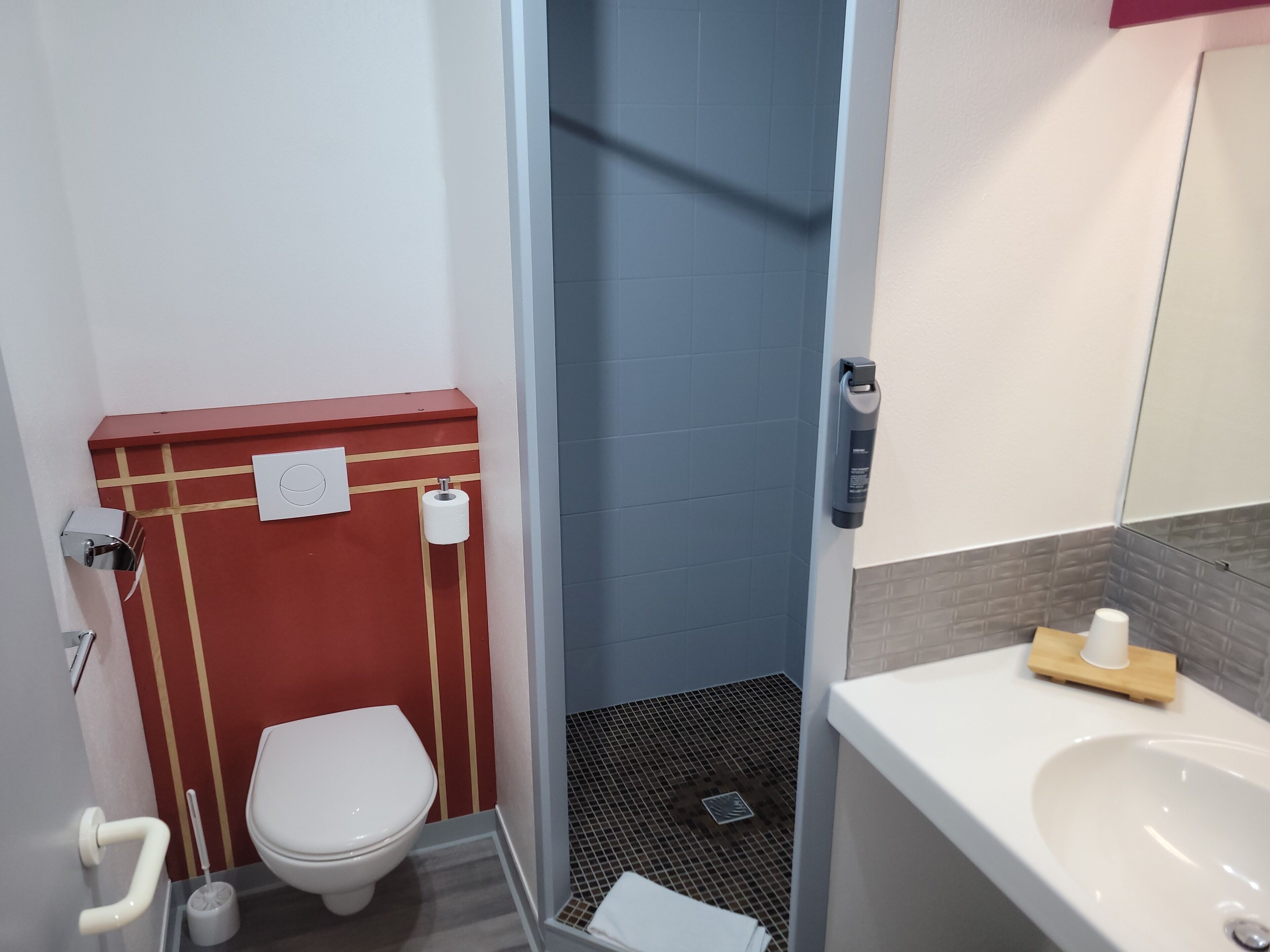 A compact bathroom with a white toilet, a red sink vanity, a shower area, and a small mirror.