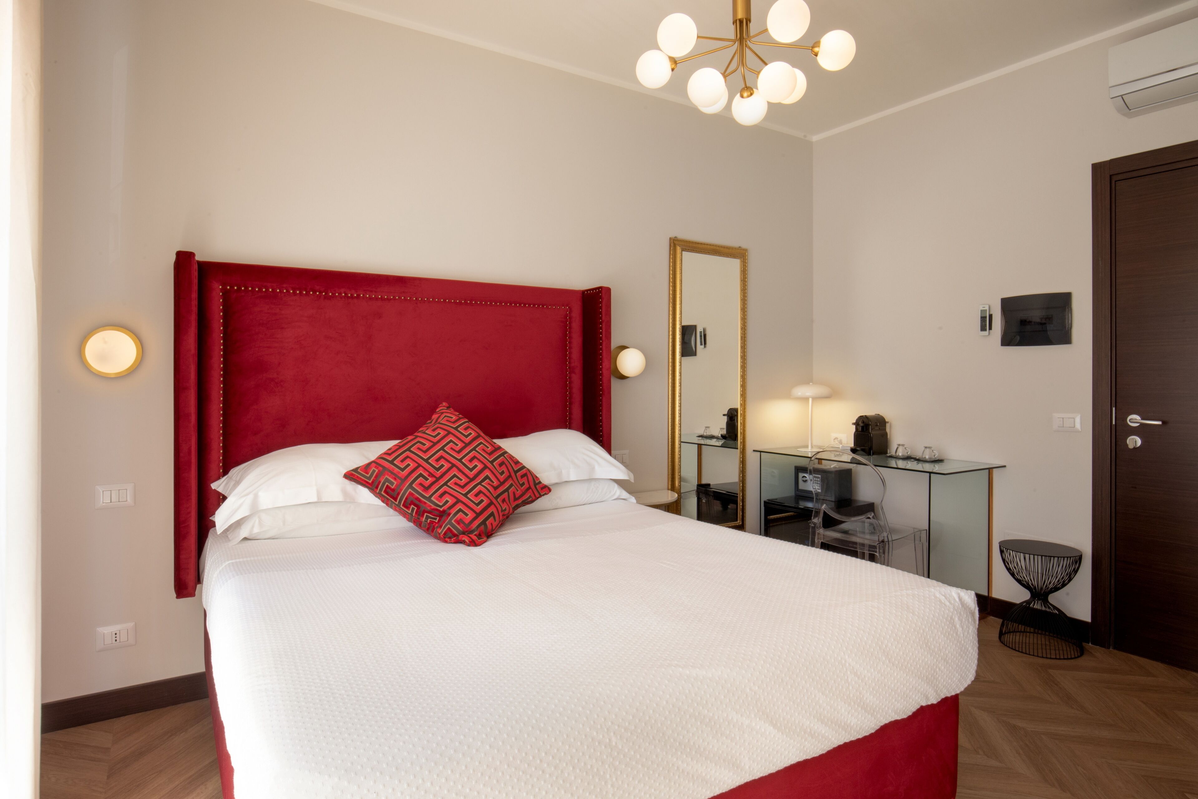 A neatly made bed with a red headboard, a bedside table with a lamp, and a mirror on the wall.