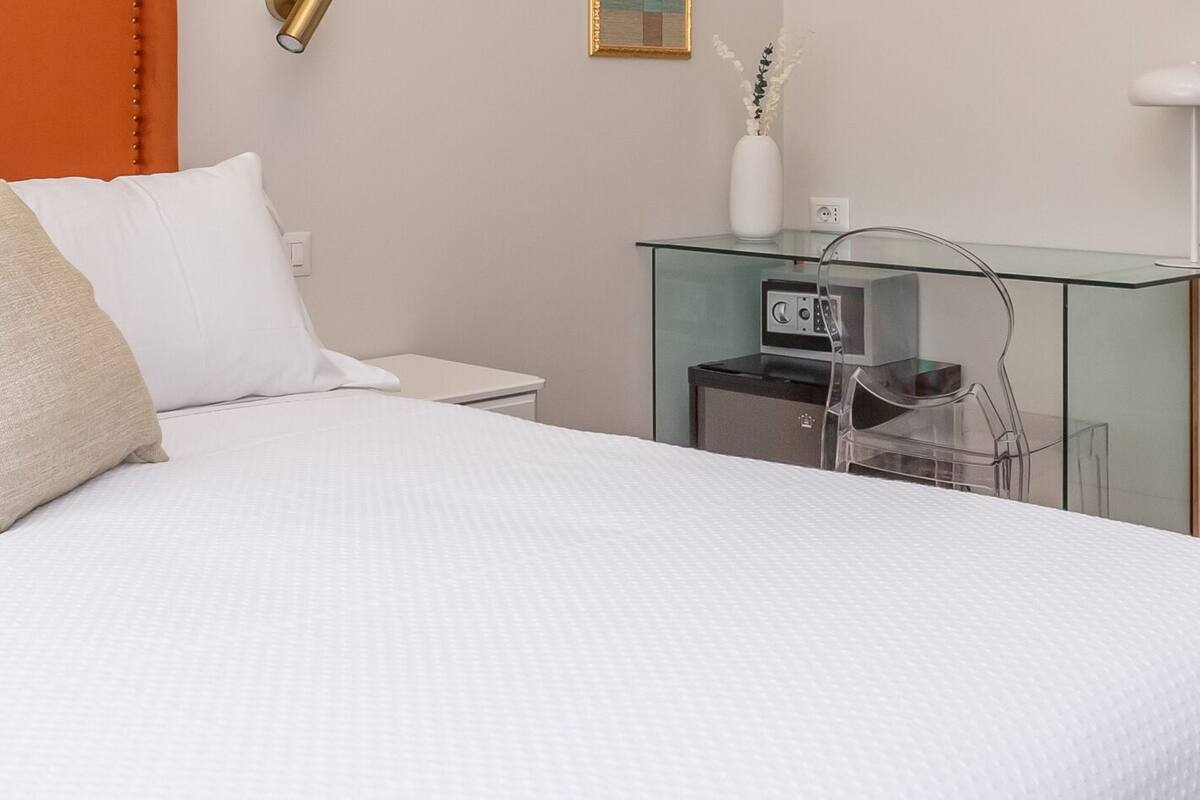 A neatly made bed with white bedding and decorative pillows, a glass side table with a lamp, and a wooden door.