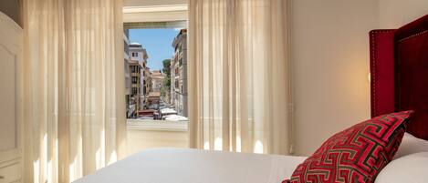 Een hotelkamer met een bed, een rood kussen met patroon en uitzicht op een straat door dunne gordijnen.