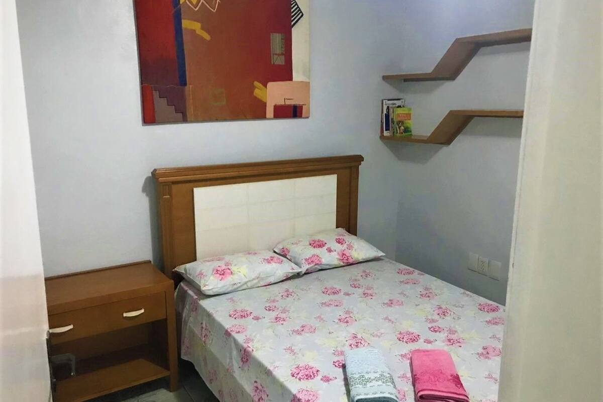 Un dormitorio pequeño y ordenado con una cama individual, una mesita de noche, un escritorio de madera y un estante en la pared.