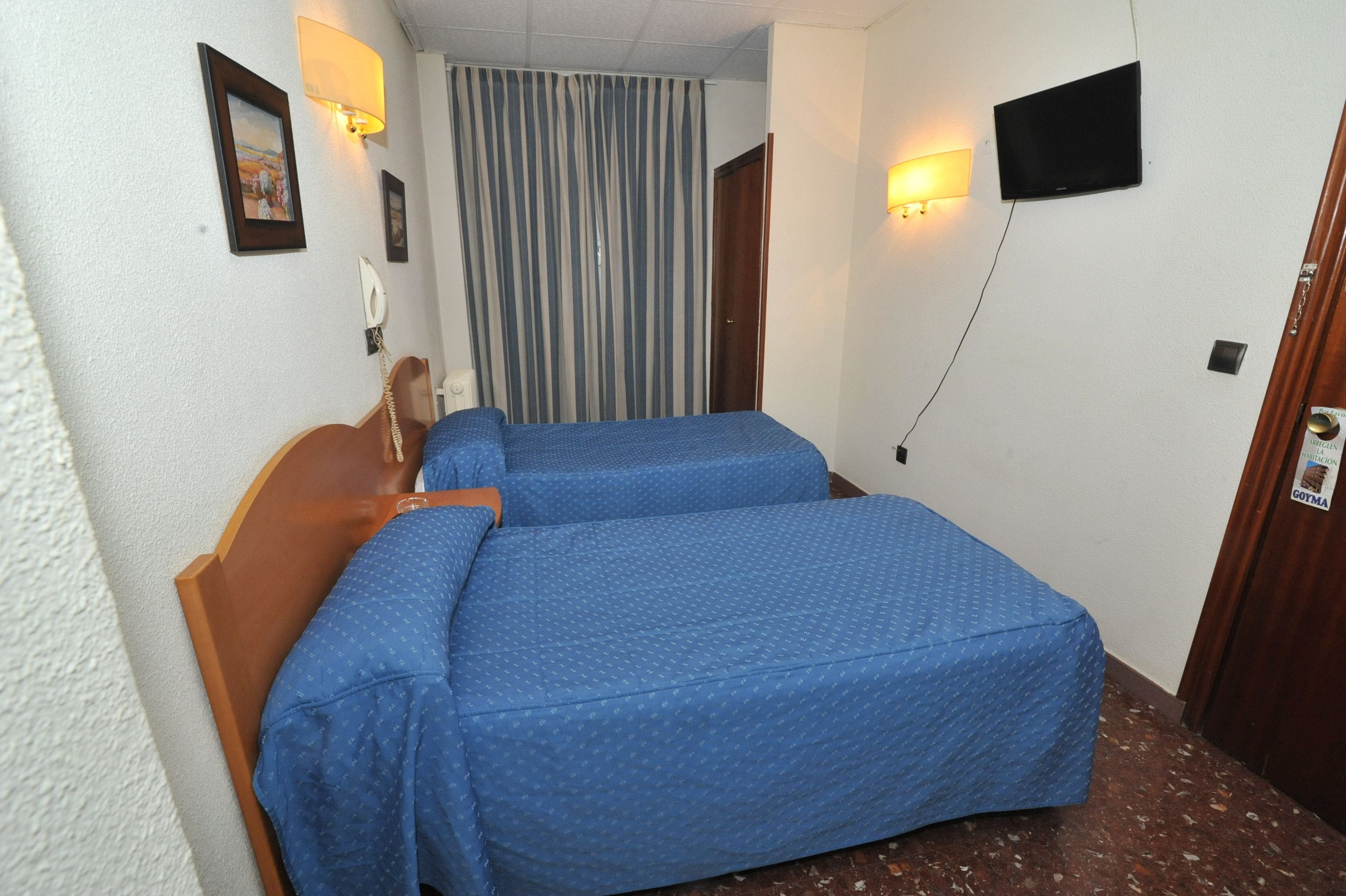Habitación de hotel con dos camas individuales, un cabecero de madera, una cortina, un televisor montado y un teléfono en la pared.