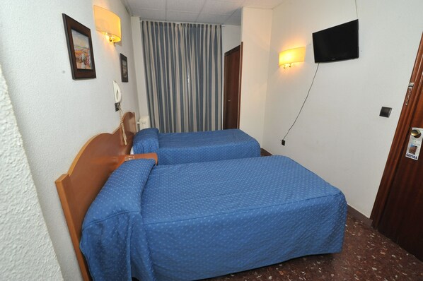 A hotel room with two single beds, a wooden headboard, a curtain, a mounted TV, and a wall-mounted phone. - Hostal Goyma I (San Fernando de Henares)