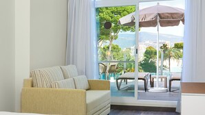 A hotel room with a bed, a sofa, a balcony with a table and chairs, and a view of greenery and buildings.
