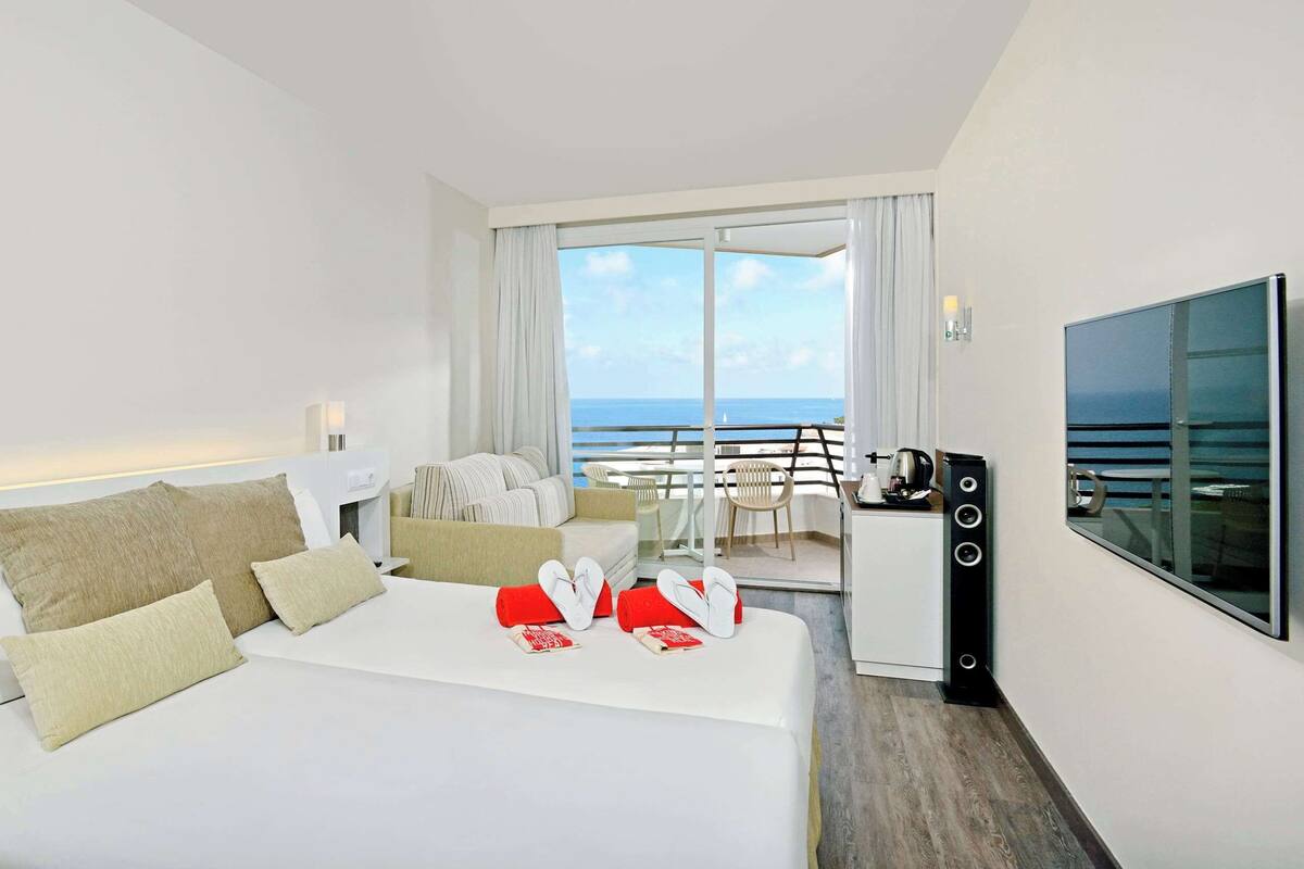 Een hotelkamer met een bed, bureau, stoel, televisie en een balkon met uitzicht op zee.