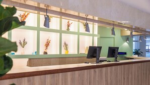 A reception desk with two monitors, a green wall with plant decorations, and hanging pendant lights.