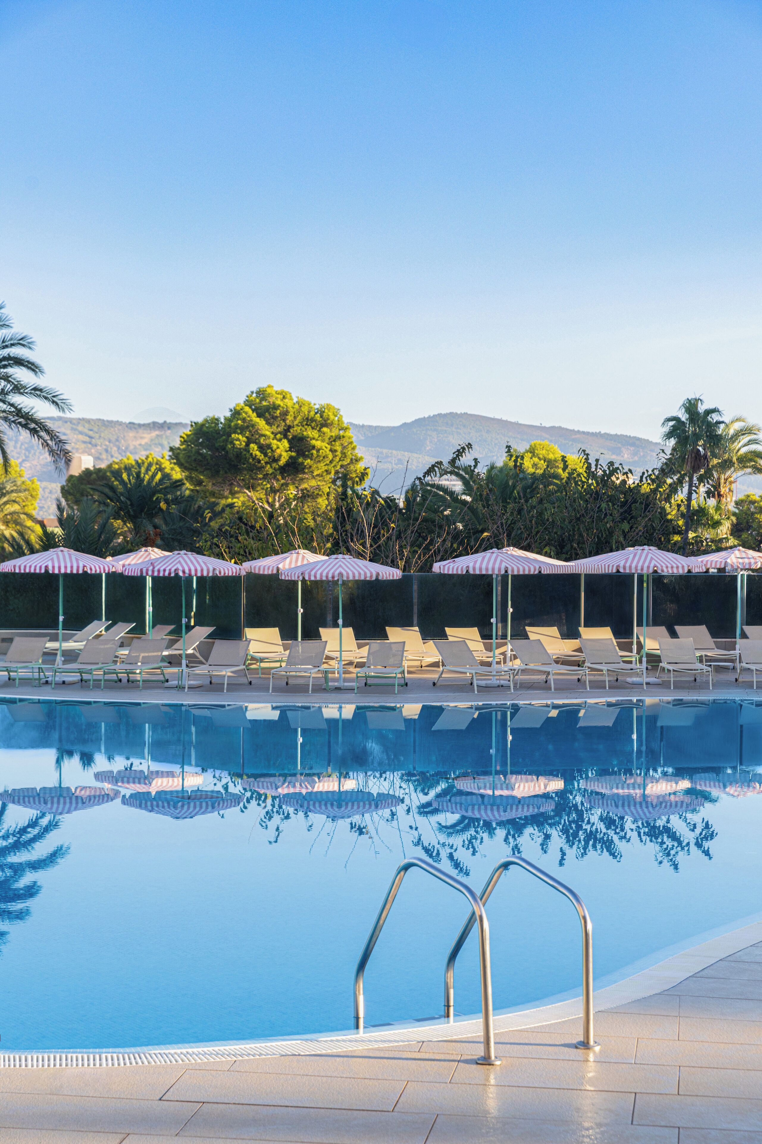 A pool with lounge chairs and striped umbrellas, surrounded by trees and a view of hills.