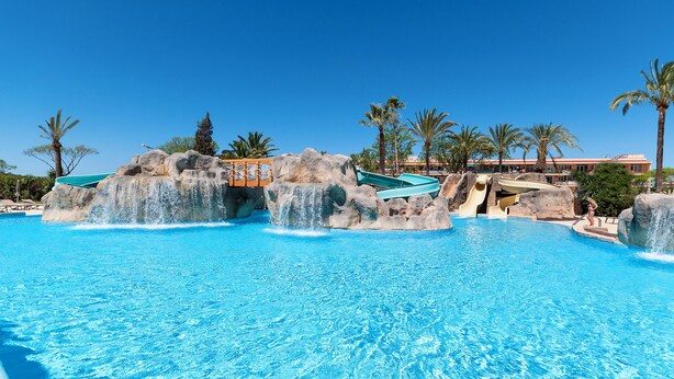 A large outdoor swimming pool with rock formations and slides, surrounded by palm trees and a clear blue sky.