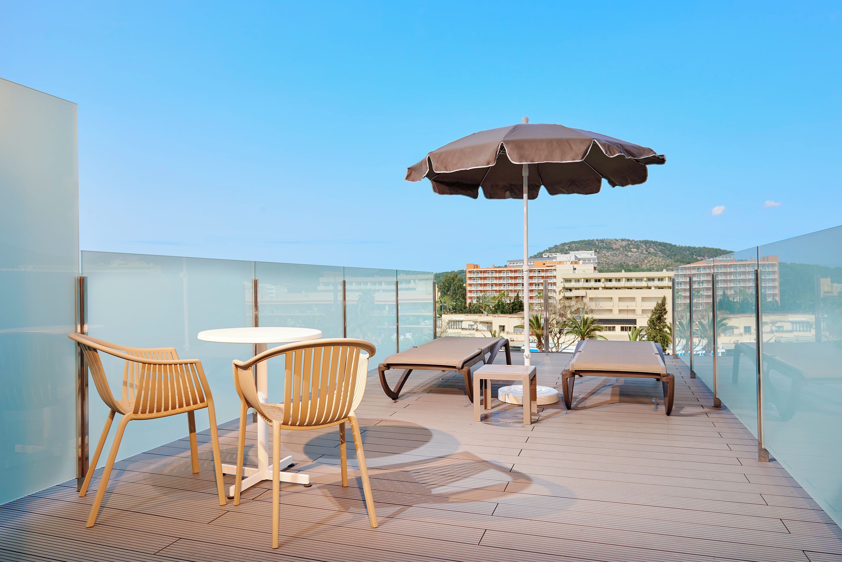 Una terraza en la azotea con barandilla de cristal, dos sillas de madera, una mesa redonda, un sillón de descanso y un paraguas grande.