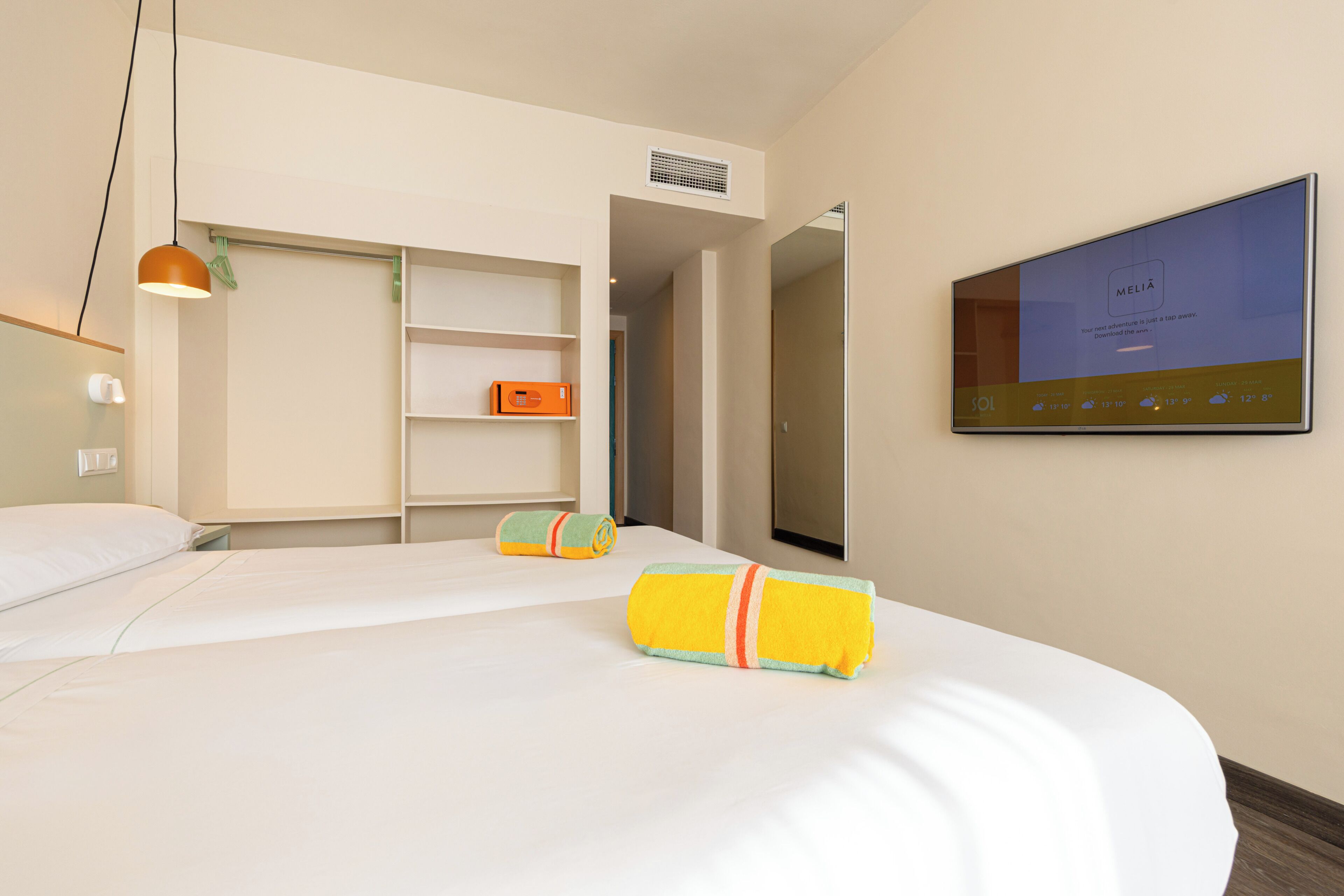 A hotel room with a bed, a television mounted on the wall, a wardrobe, and a shelf with a box.