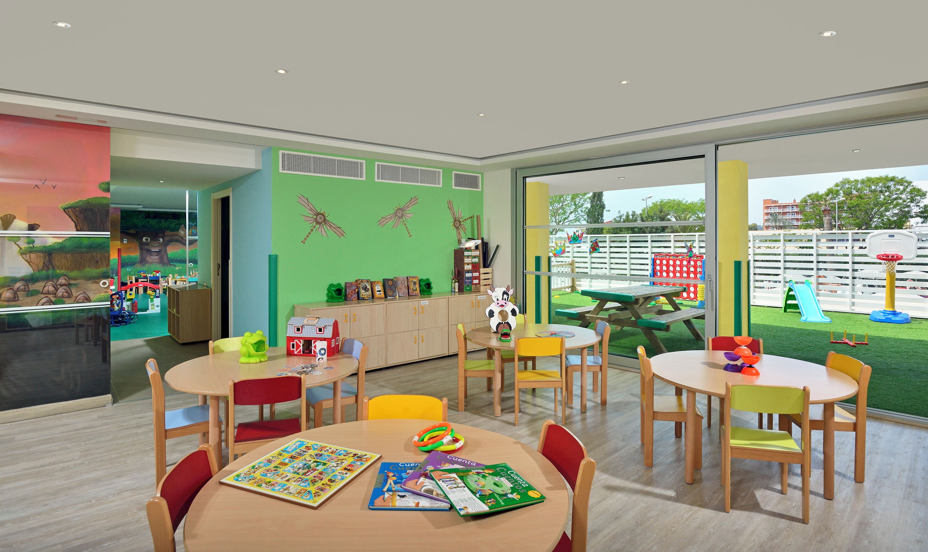 Un espacio interior luminoso para juegos, con varias mesas y sillas, un ventanal que ofrece vistas al exterior y material educativo sobre las mesas.