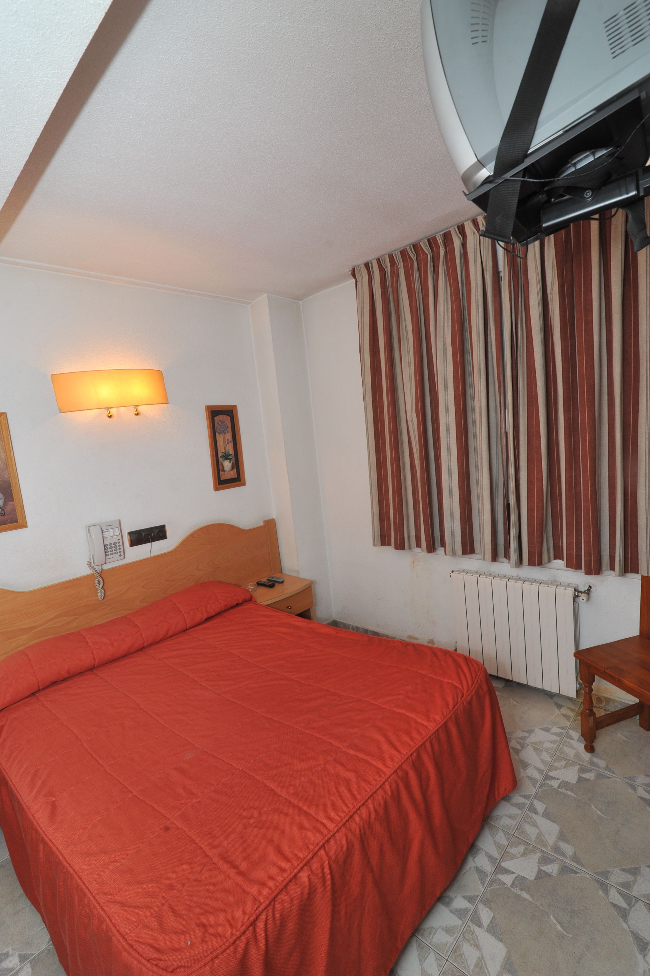 A hotel room with a large bed, a bedside table, a lamp, a telephone, a radiator, and a window with curtains.