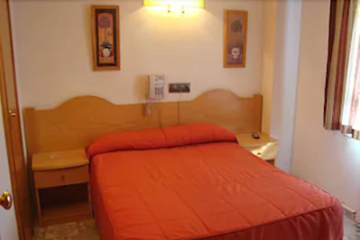 A bedroom with a wooden headboard, a single bed with a red bedspread, a bedside table with a lamp, and two framed pictures on the wall.
