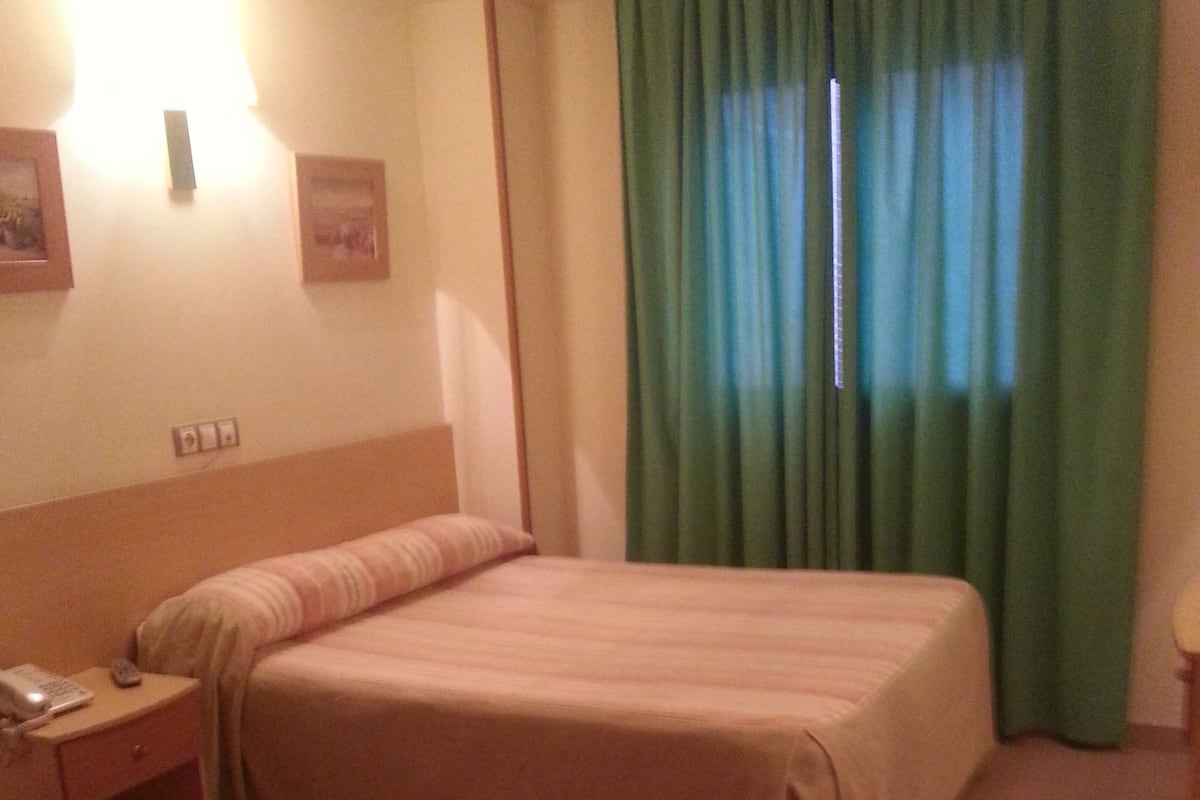 A hotel room with a bed, a nightstand, a telephone, and a window with green curtains.
