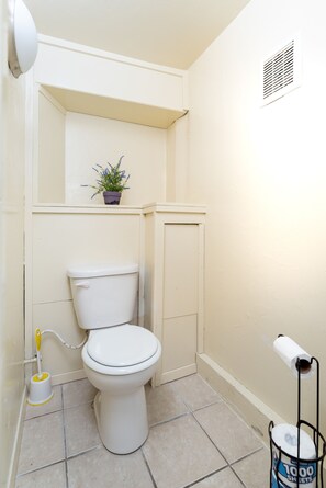 A small, clean bathroom with a toilet, a small shelf with a potted plant, a toilet paper holder, and a vent on the wall. - Bright & Spacious One Bedroom Apartment (Oakland)