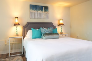 A neatly made bed with a grey headboard, two bedside tables with lamps, and a painting above the bed.
