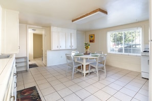 A kitchen with white cabinets, a dining table with chairs, a window with blinds, and a refrigerator.