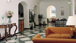 A hotel lobby with a checkered floor, a sofa, a table with a lamp, and potted plants. - Grand Hotel Ischia Lido (Ischia)