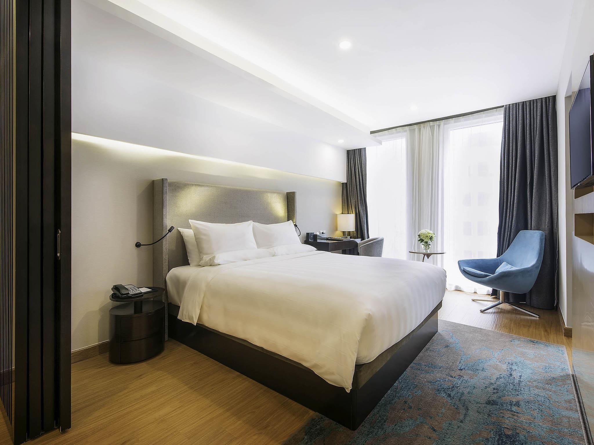 A modern hotel room with a large bed, bedside table, chair, and a TV.