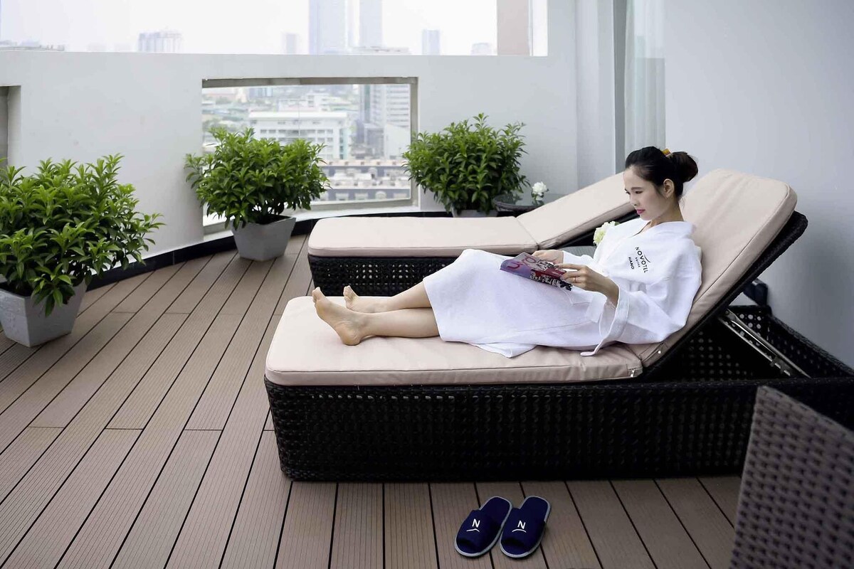 Uma pessoa relaxando em uma espreguiçadeira em uma varanda, com vista para a cidade e cercada por plantas em vasos.