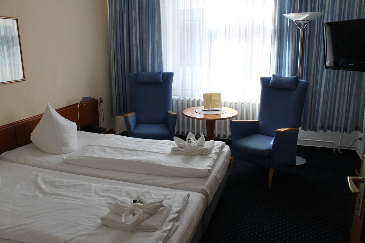 A hotel room with two beds, a round table, two blue armchairs, a TV, and a window with curtains.