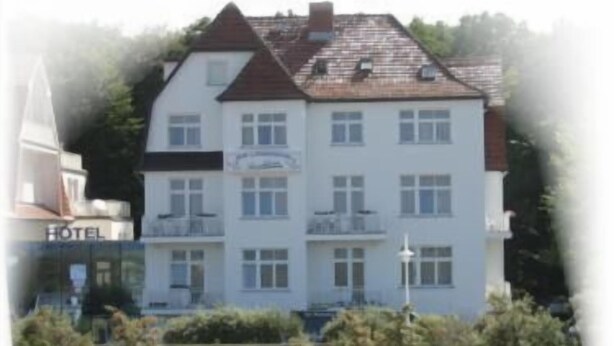 Un hotel de varios pisos con fachada blanca y techo oscuro, ubicado cerca de una playa con una torre de salvavidas y sillas de playa.