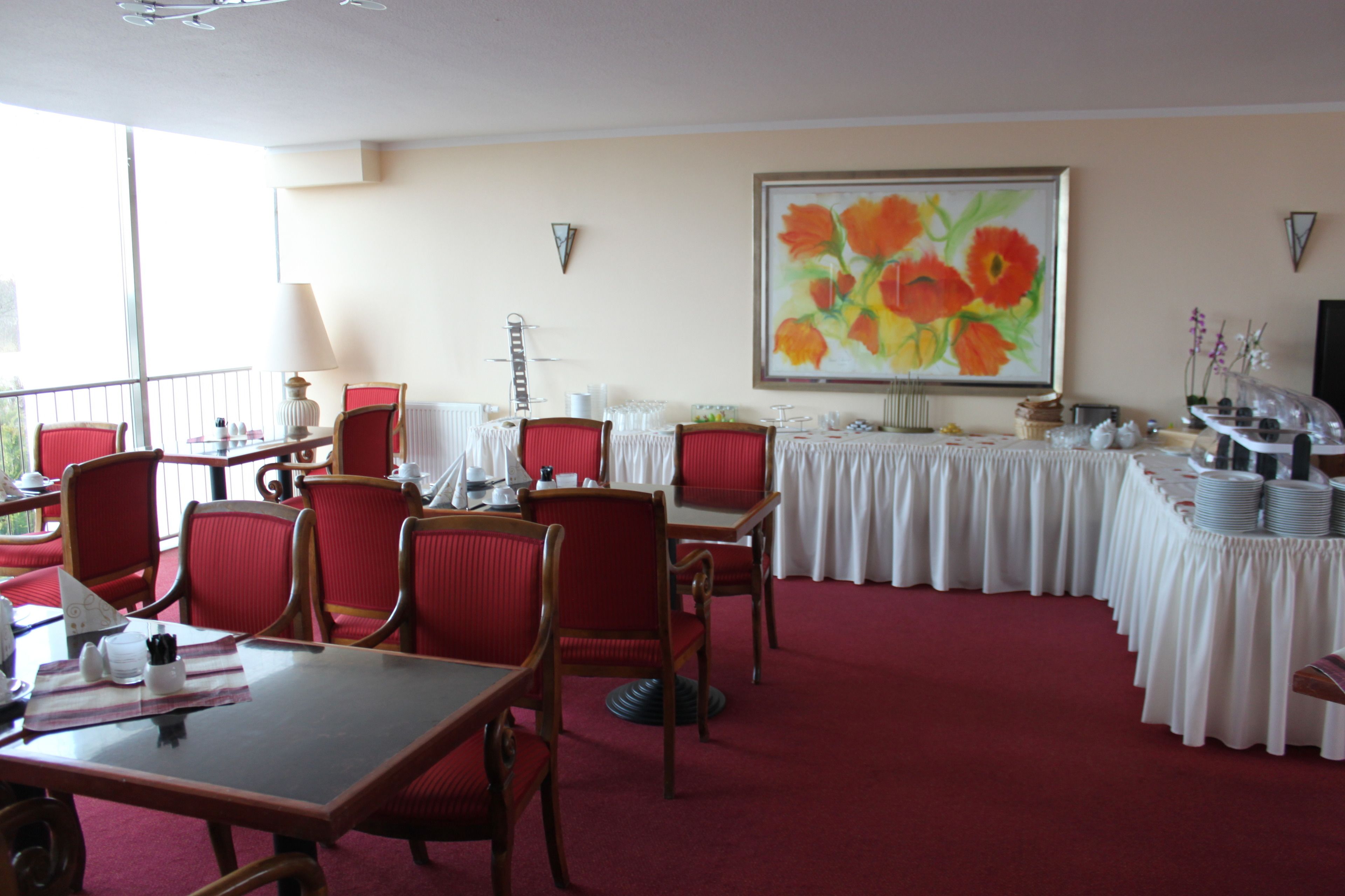 Ein Hotelrestaurant mit roten Kunstledersesseln, Tischen mit weißen Tischdecken und einem großen Blumenbild an der Wand.