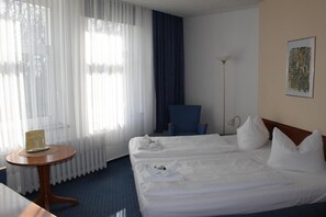 Une chambre d’hôtel avec deux lits, une petite table ronde, une chaise, un lampadaire et une fenêtre avec des rideaux légers.