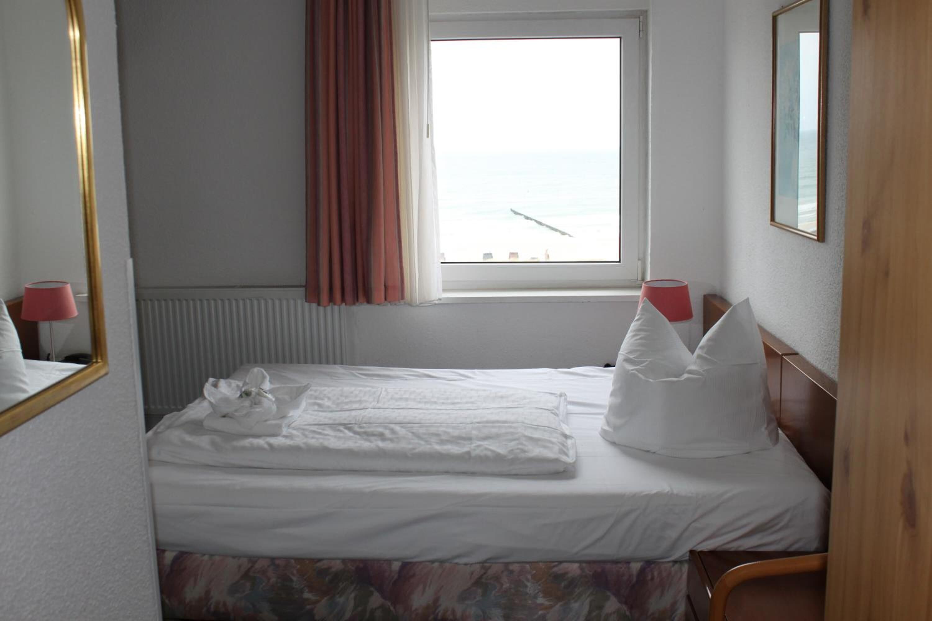 Een opgemaakt bed met witte lakens, een raam met uitzicht op zee en een spiegel aan de muur.