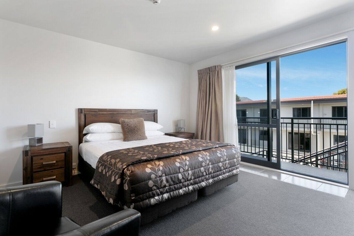 A hotel room with a large bed, a nightstand, a chair, and a balcony with a view of buildings.