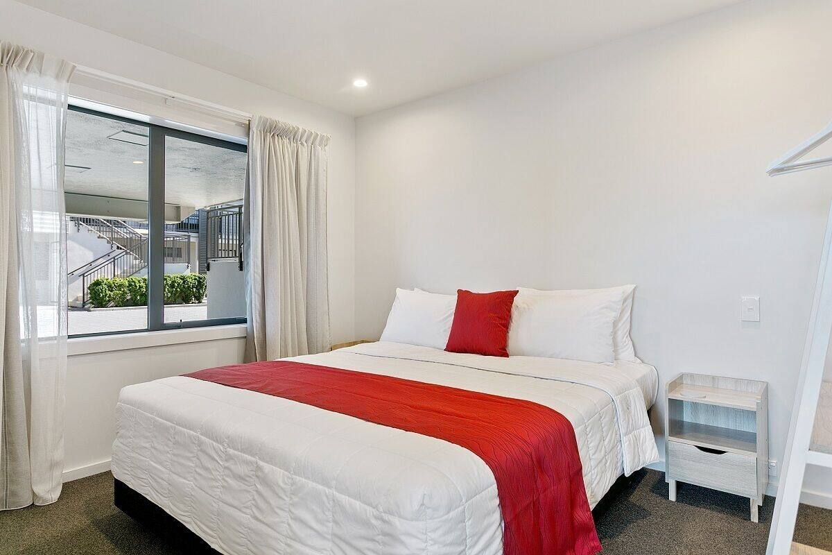 A hotel room with a large bed, a nightstand, a window with curtains, and a view of an outdoor staircase.
