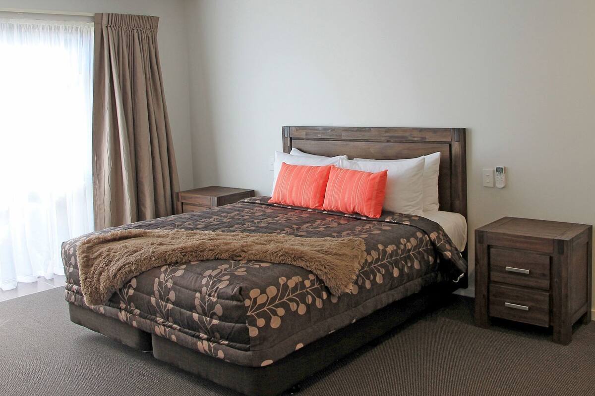 A hotel room with a bed, two nightstands, a wooden headboard, a window with curtains, and a carpeted floor.