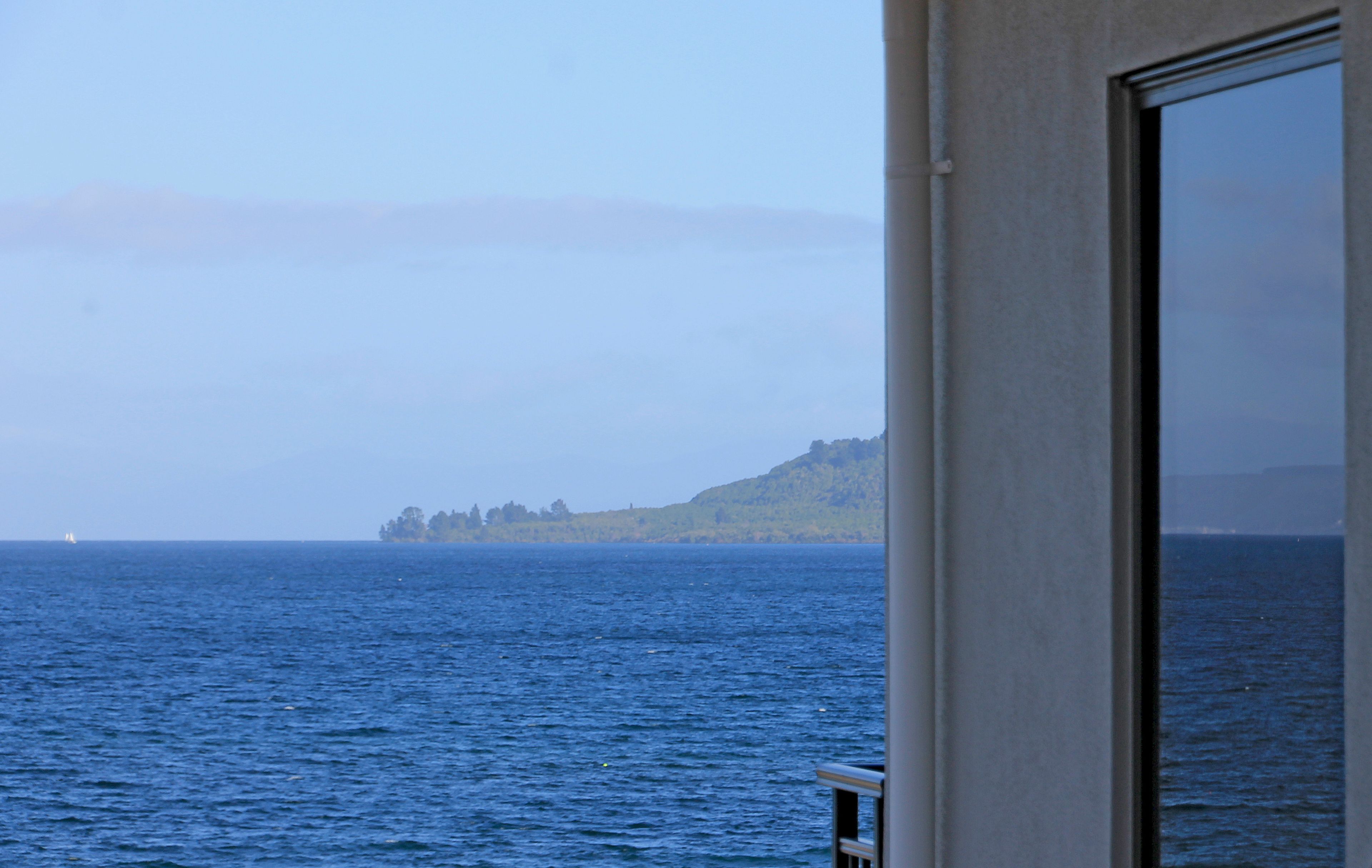 A view of the ocean from a window with a view of a distant landmass and a sailboat.